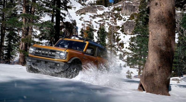 An orange 2021 Ford Bronco 2dr is off-roading in the snow in the woods.