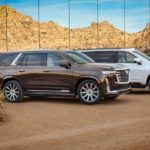 A brown and a white 2021 Cadillac Escalade are parked in front of a reflective building in the desert.