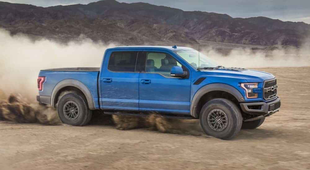 A blue 2020 Ford Raptor is kicking up dust while off-roading on dirt in front of a mountain.