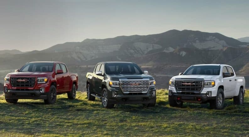 A red, a black, and a white 2021 GMC Canyon are parked on grass in front of mountains.
