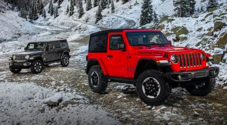 A red 2018 used Jeep Wrangler is parked in front of a grey Unlimited on a snowy mountain trail.