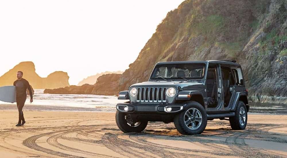 A grey 2018 used Jeep Wrangler Unlimited with no doors is on the beach next to a surfer.