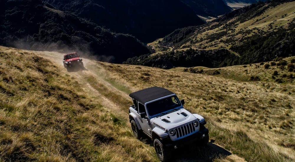 A white and a red 2018 used Jeep Wrangler are driving on a dirt path through hills.