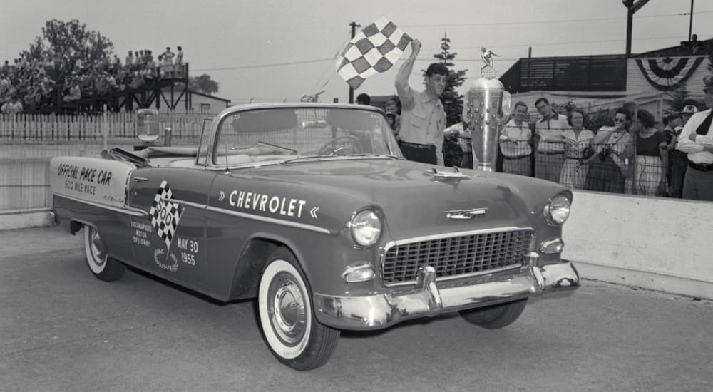 A 1955 Chevrolet BelAir pace car is parked next to a crowd with a man waving a checkered flag.