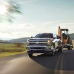 A black 2016 Chevrolet Silverado is towing a yellow front and backhoe loader down the road.