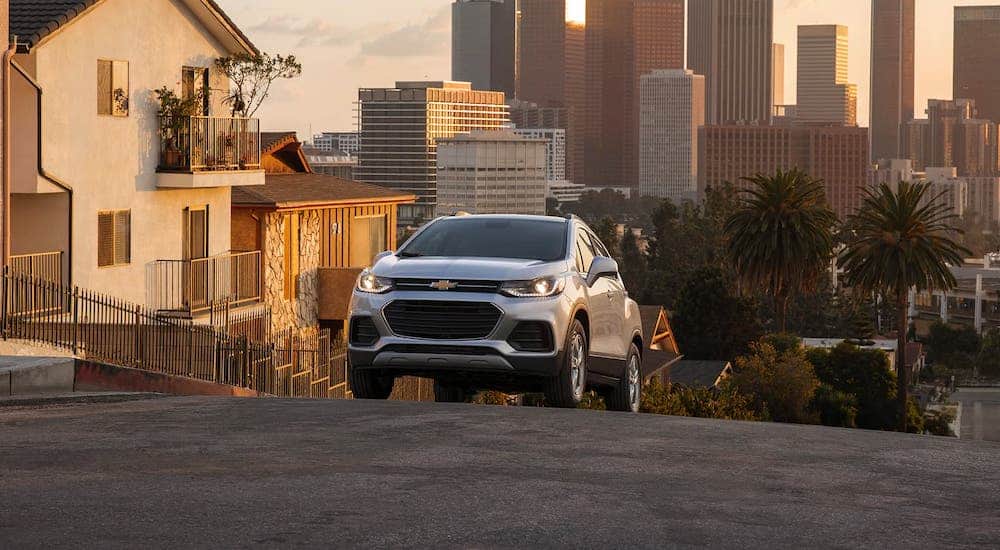 A silver 2021 Chevy Trax is shown from the front while driving up a hill and a city is shown in the distance.