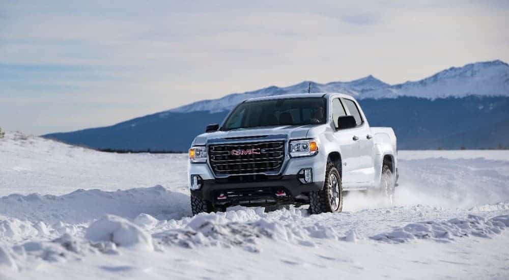 A white 2021 GMC Canyon AT4 is driving through the snow with mountains looming in the distance.