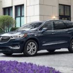 A dark blue 2020 Buick Enclave Avenir from a Buick dealer is parked outside a grey building with purple flowers.