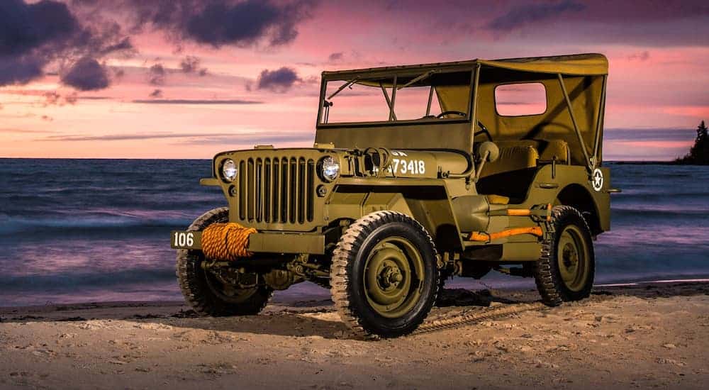 A green 1940s Jeep Willys MB is parked on a beach at sunset.