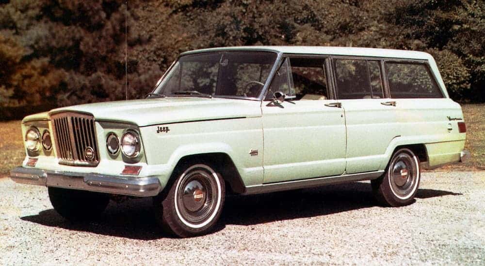 A light green 1960s Jeep Wagoneer is parked in front of trees.