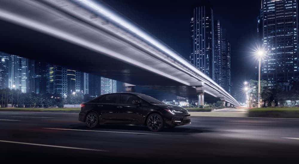 A black 2021 Toyota Corolla SE Nightshade Edition is driving under a bridge through the city at night.
