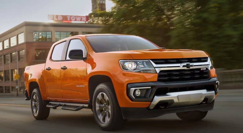 An orange 2021 Chevy Colorado is driving past buildings and trees after winning the 2021 Chevy Colorado vs 2021 Jeep Gladiator comparison.