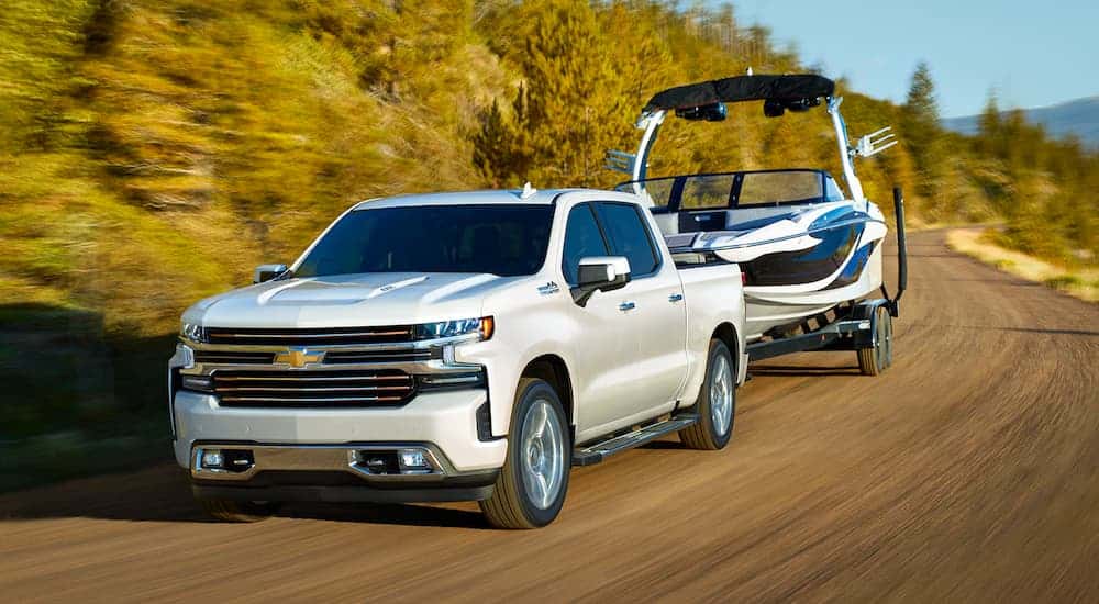 A white 2021 Chevy Silverado 1500 is towing a boat down a mountain road with trees.