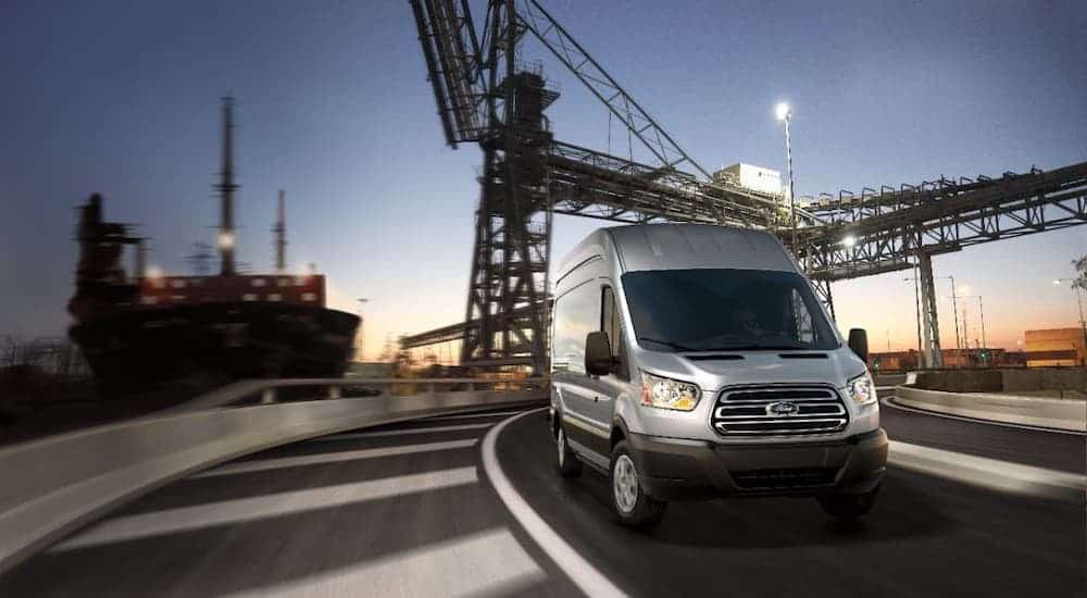 A silver 2021 Ford Commercial Transit Van is driving past a metal scaffolding at dusk.