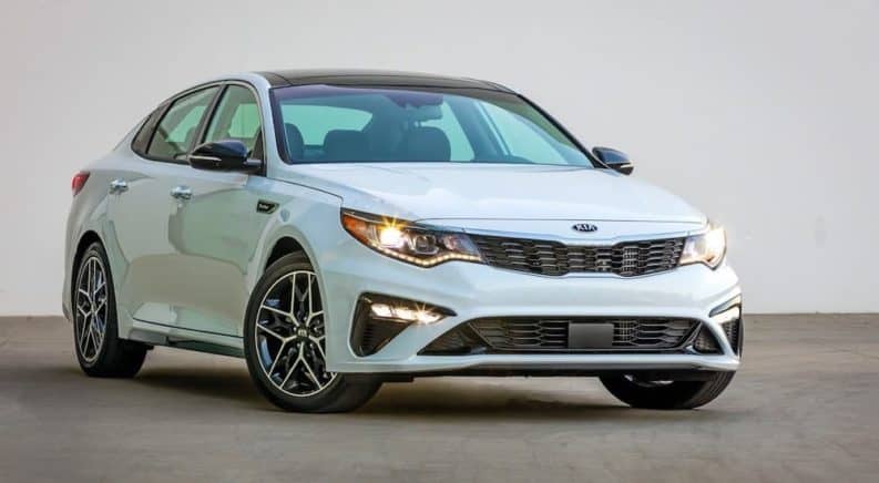 A white 2020 Kia Optima is shown from the front, angled right, with its lights illuminated.