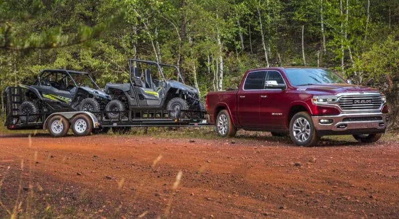 A dark red 2021 Ram 1500 is towing two Side-by-sides on a trailer on a dirt trail.