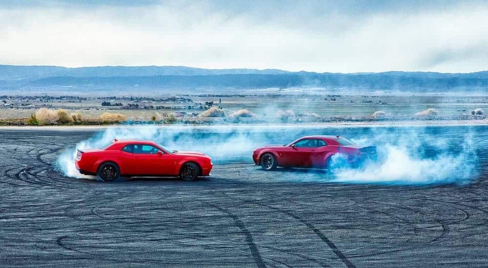 An orange and a red 2021 Dodge Challenger are doing doughnuts in an emptry parking lot.