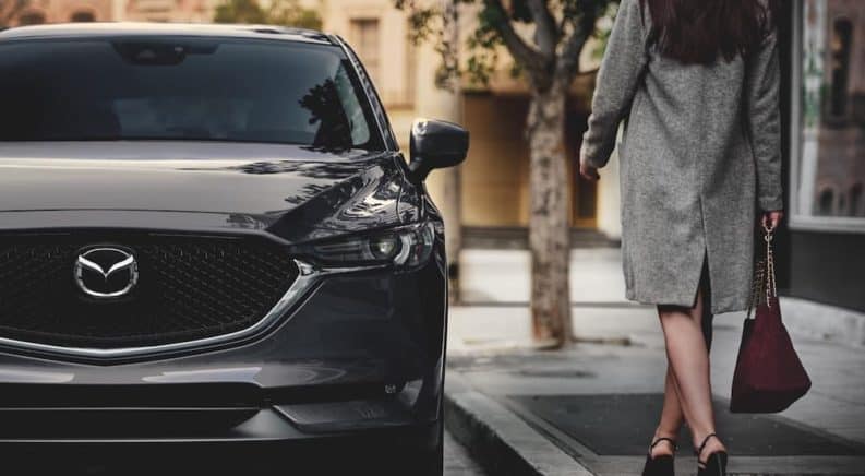 A close up is shown of the front of a dark grey 2021 Mazda CX-5 with a woman approaching it.