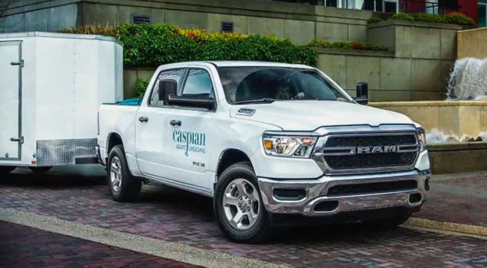 A white 2021 Ram 1500 with a landscaper's name on the door and white enclosed trailer attached is parked in front of an office building.