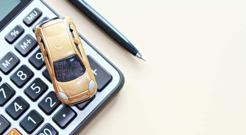 A bronze toy car is shown from a high angle parked on a calculator next to a pen.