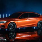 An orange 2022 Honda Civic prototype is parked in a dark warehouse in front of neon lights.
