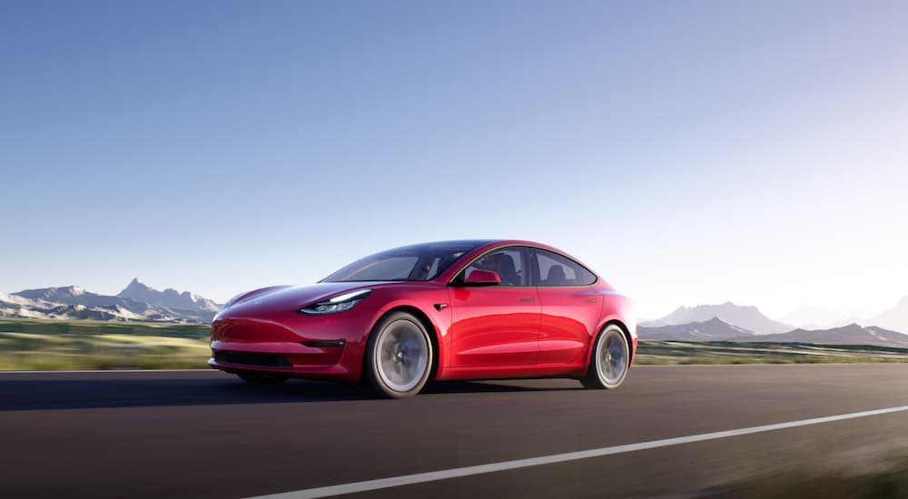 A red 2021 Tesla Model 3 is driving down the highway with blue skies in the background.