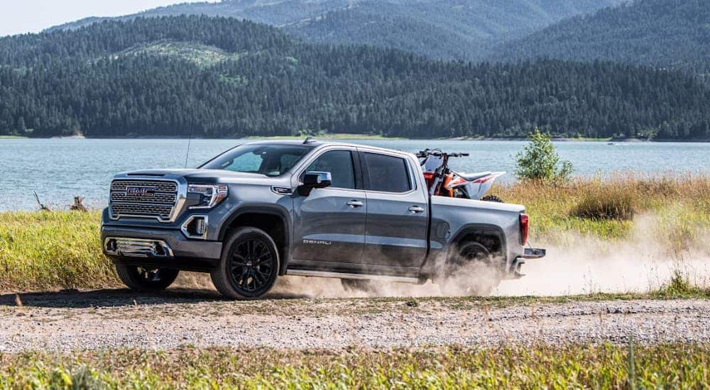 A light gray 2021 GMC Sierra Denali with a dirtbike in the bed is kicking up dirt on a dirt road in front of a lake.
