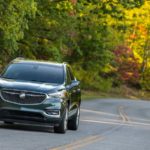 A dark grey 2019 used Buick Enclave Avenir is shown from the front driving down a heavily wooded road.