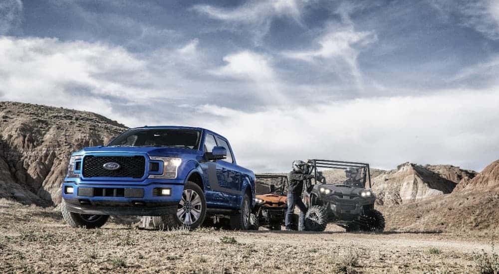 A blue 2018 used Ford F-150 is parked in a field with two UTVs behind it.