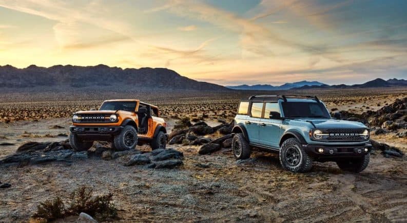 A yellow 2021 Ford Bronco 2-door with no doors and blue 2021 Ford Bronco 4-door are parked on rocks in a desert.