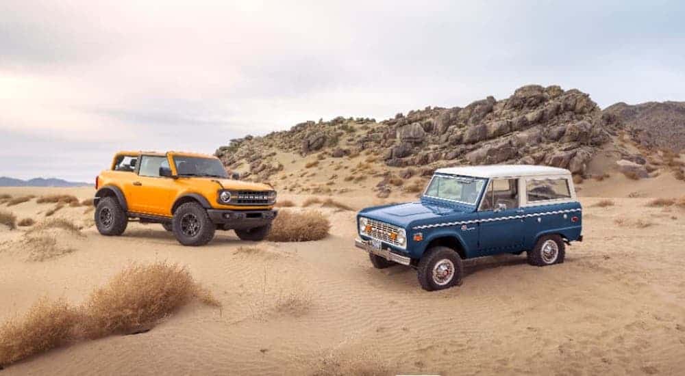 A yellow 2021 Ford Bronco 2-door with no roof is parked next to a 1960's Bronco on sand.