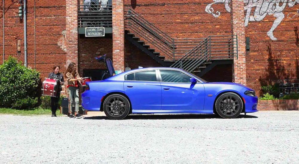 A band is loading a drum set into the trunk of a blue 2021 Dodge Charger that is parked in front of a red brick building.