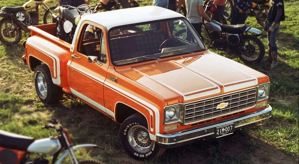 An orange and white 1976 Chevy C10 Step side is parked in a field surrounds by dirt bikes.