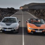 A silver and an orange 2019 BMW i8 are driving next to each other down an empty road.