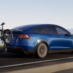 A blue 2021 Tesla Model X with bikes on the back, not something you would easily find among used cars, is driving on a highway past a body of water.