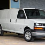 A white 2021 Chevy Express is shown parked in front of a warehouse with the driver loading cardboard boxes.