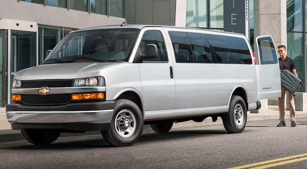 A silver 2021 Chevy Express is shown from the side, at an airport, with luggage being loaded into the back.