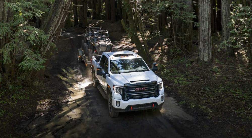 A white 2021 GMC Sierra 2500HD AT4 is shown from a high angle towing a trailer on a dirt road in the woods.