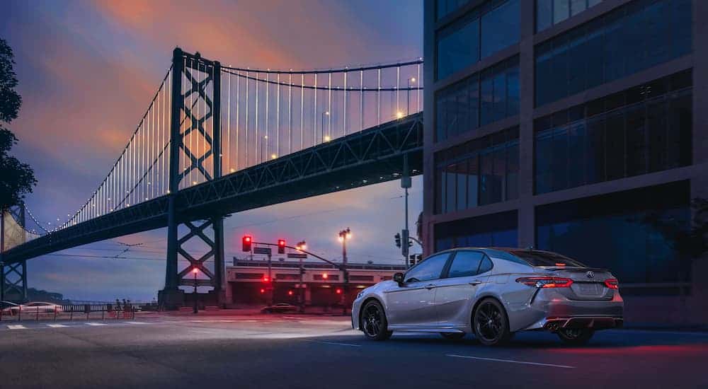 A popular used car, a silver 2018 Toyota Camry, is driving through the city past a bridge.