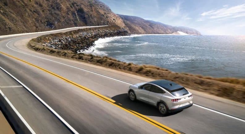 A white 2021 Ford Mustang Mach-E is shown from a high angle driving on a winding coastal highway.