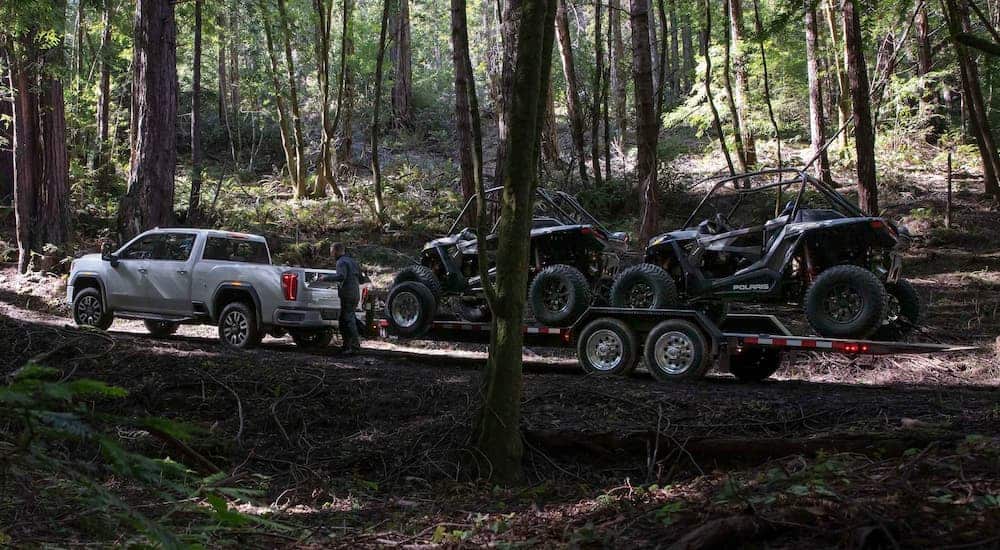 A white 2021 GMC Sierra 2500HD AT4 is shown from the side towing UTVs in the woods.