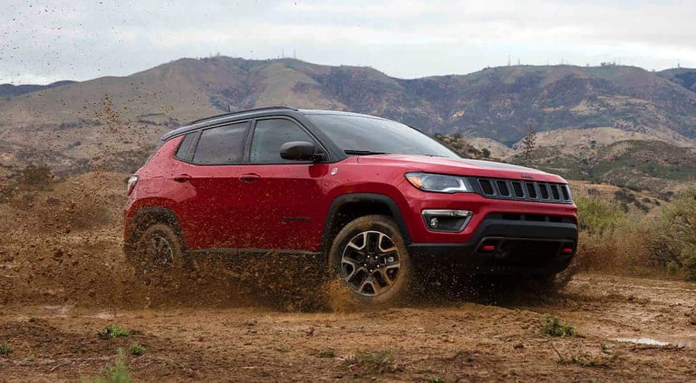 A dark red 2021 Jeep Compass Trailhawk is off-roading in mud in front of mountains after leaving a Jeep Compass dealer.