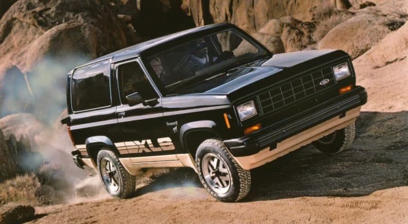 A popular used car for sale near you, a black and gold 1980s Ford Bronco II XLS, is driving on a dirt road.