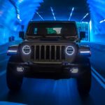 A silver 2021 Jeep Wrangler 4xe is shown from the front driving though a tunnel illuminated in blue.