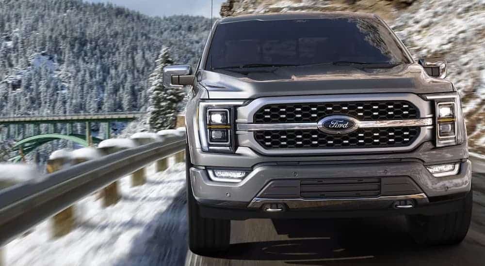 A gray 2021 Ford F-150 is shown from the front driving through the mountains past a green bridge.