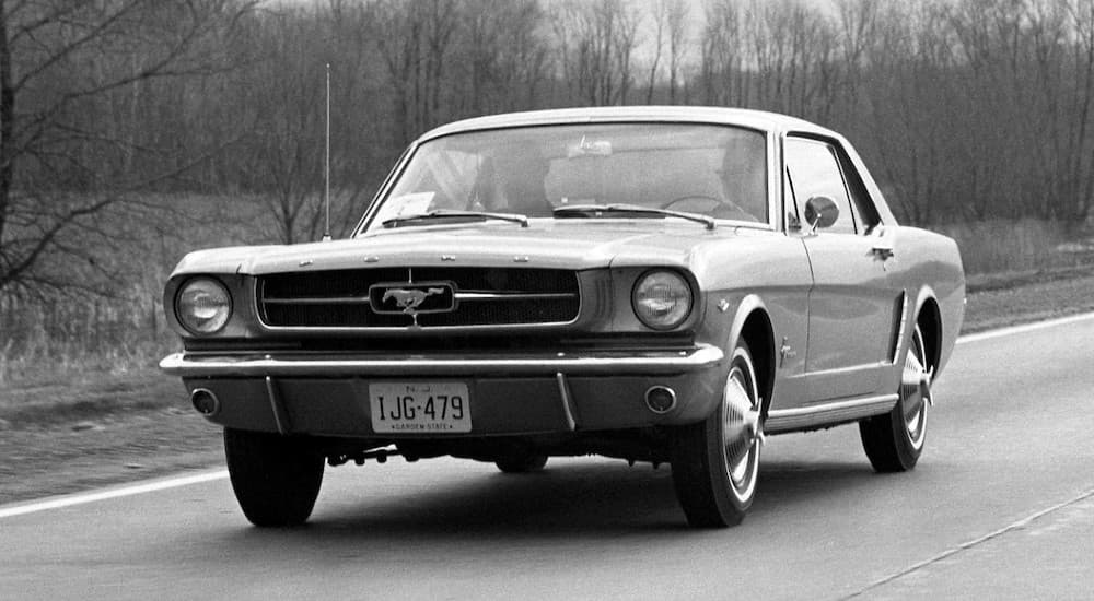 A classic famous car, a 1964 Ford Mustang, is shown in black and white driving down the road.