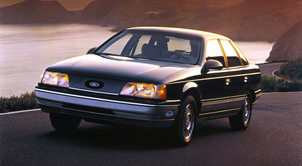 A blue 1986 Ford Taurus is shown from the front with a lake in the background after visiting a Ford dealership.