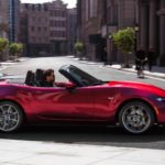 A red 2021 Mazda MX-5 Miata is shown from the side driving through the city after leaving a Mazda dealer.