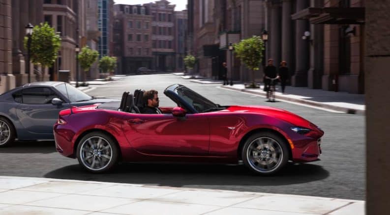 A red 2021 Mazda MX-5 Miata is shown from the side driving through the city after leaving a Mazda dealer.