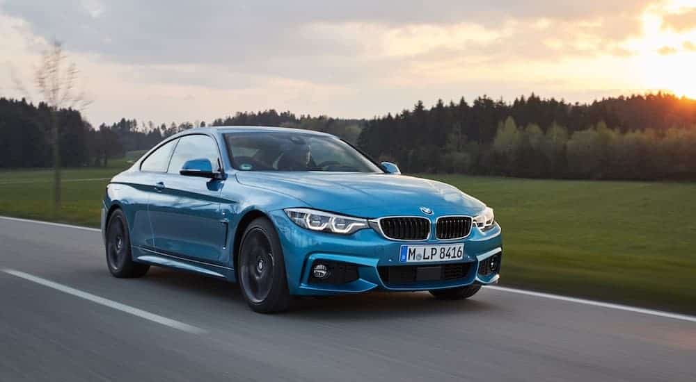 A blue 2017 BMW 4 Series coupe is driving past a field with the sun setting over trees.